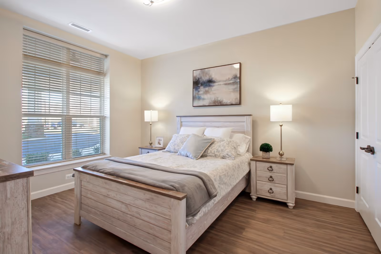 A bright and cozy bedroom with a large window covered by blinds, a wooden bed with white and gray bedding, two matching nightstands each with a lamp, a small plant on one nightstand, and a framed picture on the other. A landscape painting hangs above the bed, and the floor is wood.