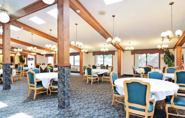 A spacious dining room with multiple round tables covered with white tablecloths, surrounded by green cushioned wooden chairs. The room features wooden beams and columns, carpeted floor, large windows with valances, and several chandeliers providing warm lighting. There are flowers on the tables and an American flag in the corner.