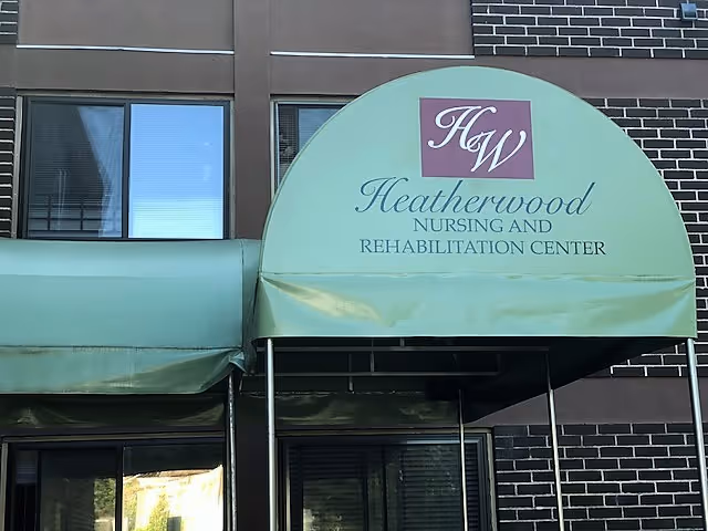 Entrance of Heatherwood Nursing and Rehabilitation Center with a green awning displaying the facility's name and logo, set against a brick and windowed building exterior.