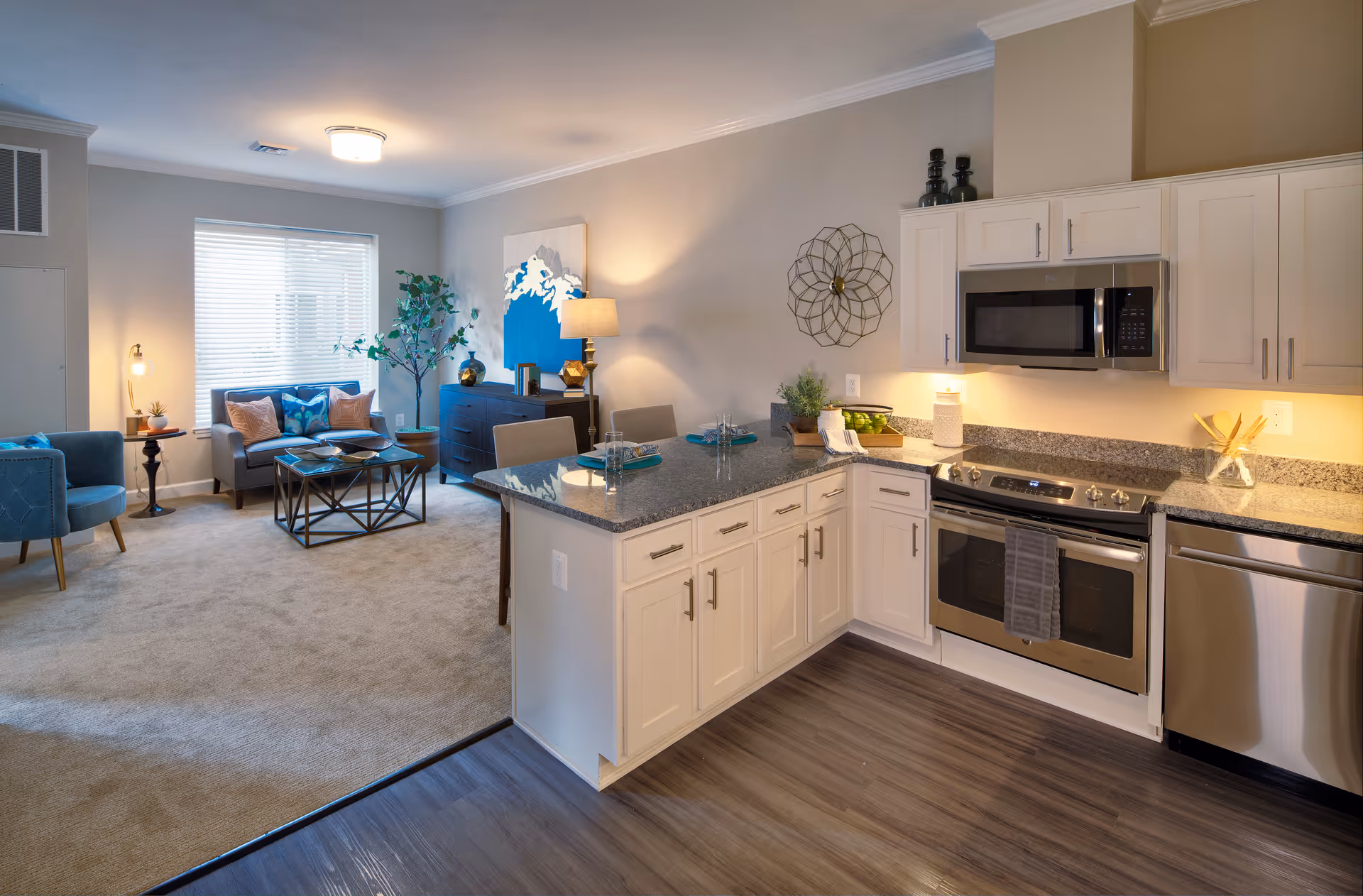 A modern open-concept living space featuring a kitchen with white cabinets, granite countertops, stainless steel appliances including a microwave, oven, and dishwasher. Adjacent to the kitchen is a cozy living room area with a beige carpet, a gray sofa with blue and pink cushions, a blue armchair, a coffee table, a side table with a lamp, and decorative plants and artwork on the walls.