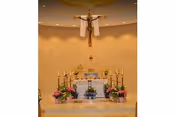 Interior view of a chapel altar with a large crucifix mounted on the wall above. The altar is decorated with candles, flowers in pots, and religious items. The background wall is beige with a subtle texture.