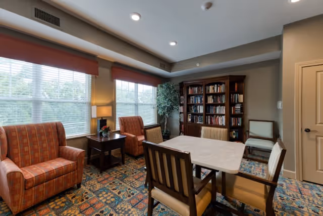 A cozy interior room with a rectangular table surrounded by six chairs, two orange armchairs near large windows with blinds, a side table with a lamp and a small plant, a tall bookshelf filled with books, and a potted plant in the corner.