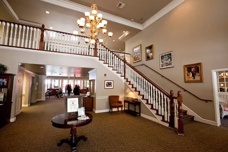 Interior view of a senior living facility lobby featuring a carpeted floor, a wooden staircase with white balusters and dark handrails, a chandelier hanging from the ceiling, framed pictures on the wall, a small table with a lamp and chair underneath the stairs, and a round table with framed documents in the center. In the background, several people are visible near a seating area with large windows letting in natural light.