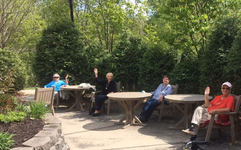 Four elderly people sitting at outdoor tables in a garden area, waving and smiling. The area is surrounded by green bushes and trees, with a stone border along a garden bed.