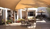 Outdoor patio area at Freedom Plaza Care Center with tables and chairs under shade sails, surrounded by plants and flowers, with a water fountain and building windows in the background.