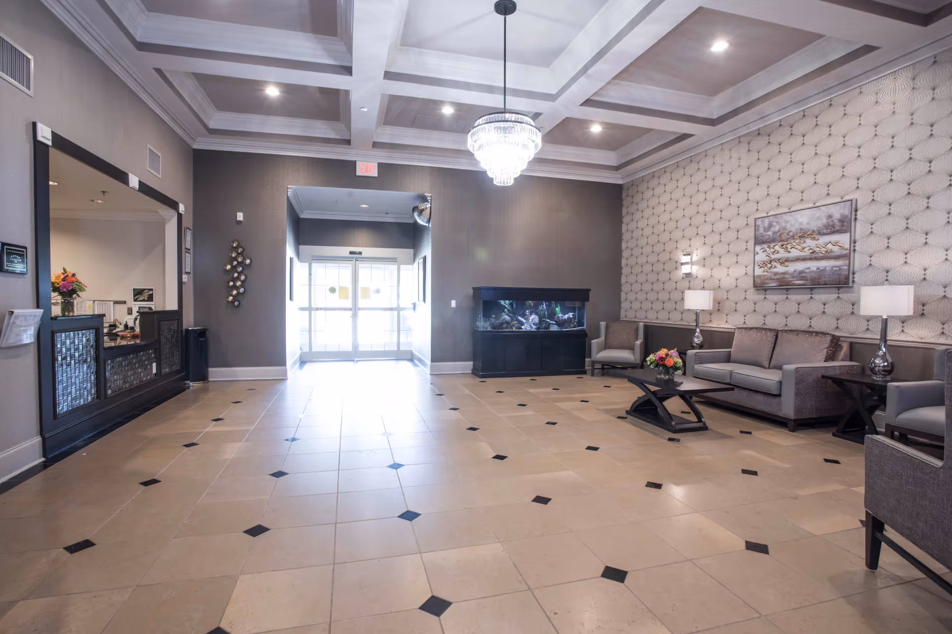 A spacious and well-lit lobby area with beige tiled flooring featuring black diamond accents. The ceiling has a coffered design with recessed lighting and a central chandelier. On the right side, there is a seating area with a gray sofa, two armchairs, a coffee table with a flower arrangement, two side tables with lamps, and a large fish tank against the wall. The far end shows glass double doors leading outside. The left side has a reception window with a counter and some flowers on it.