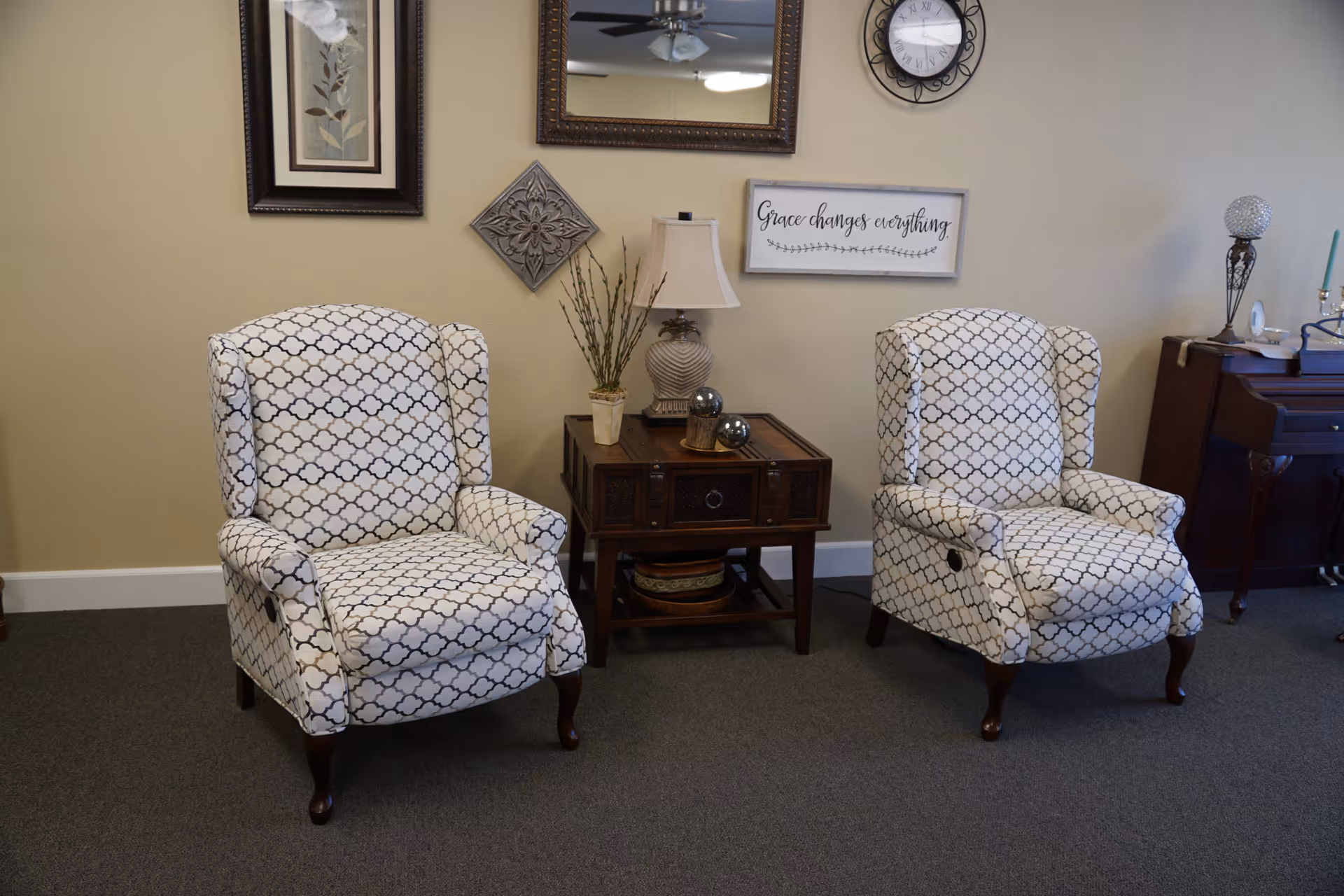 Two patterned armchairs flank a wooden side table with a lamp and wall decor in a seating area.