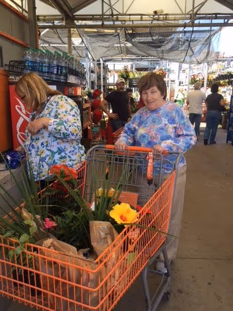 Two women shopping at a garden center with an orange shopping cart filled with plants and flowers. One woman is pushing the cart and smiling, while the other is looking at items on a shelf. Other shoppers and plants are visible in the background under a covered outdoor area.