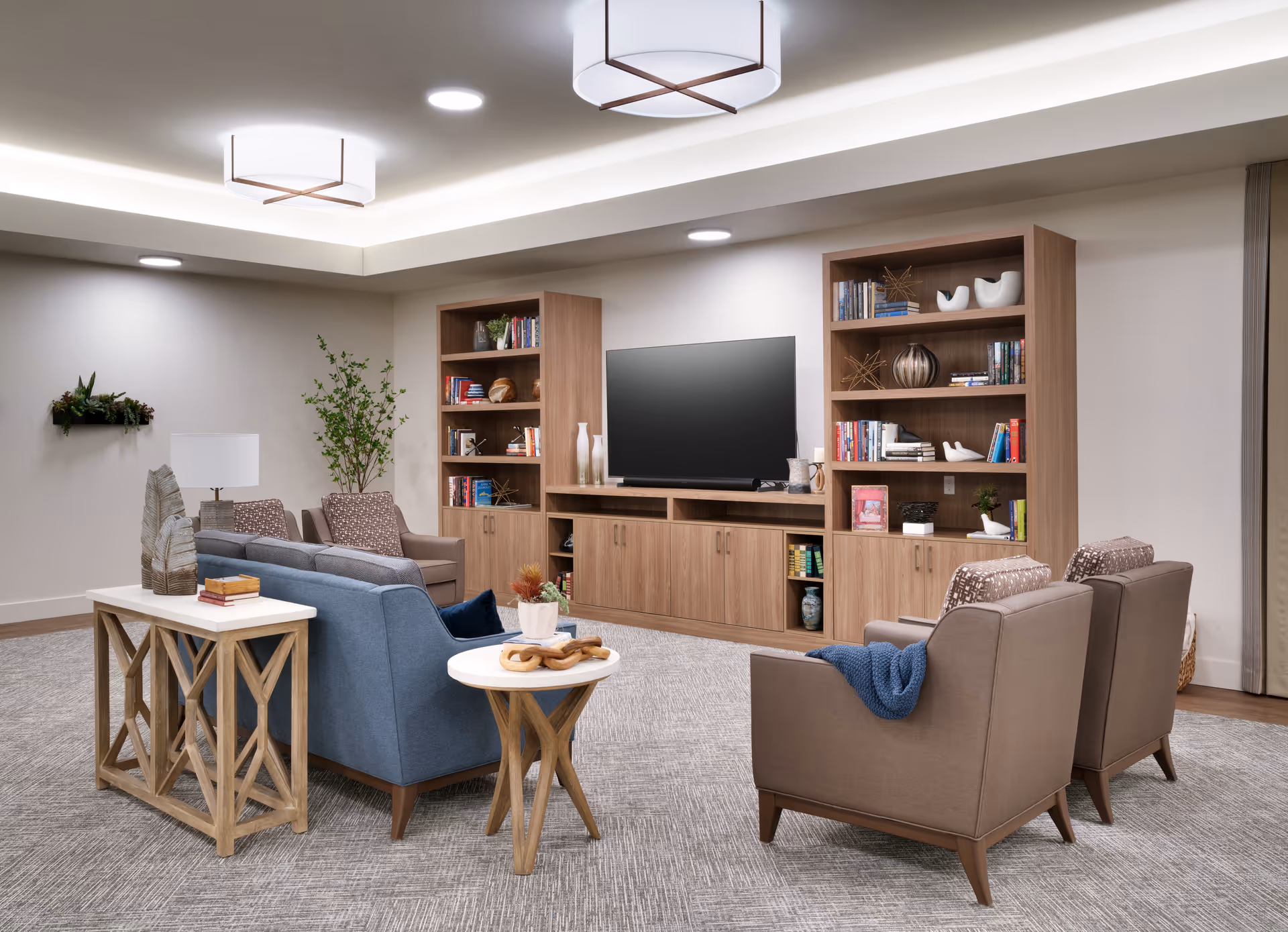 A cozy senior living room featuring a blue sofa, two brown armchairs with a blue throw, a wooden side table with decorative items, and a large wooden entertainment center with a flat-screen TV and bookshelves filled with books and decor. The room has soft lighting from ceiling fixtures and a neutral color palette with beige walls and carpet.
