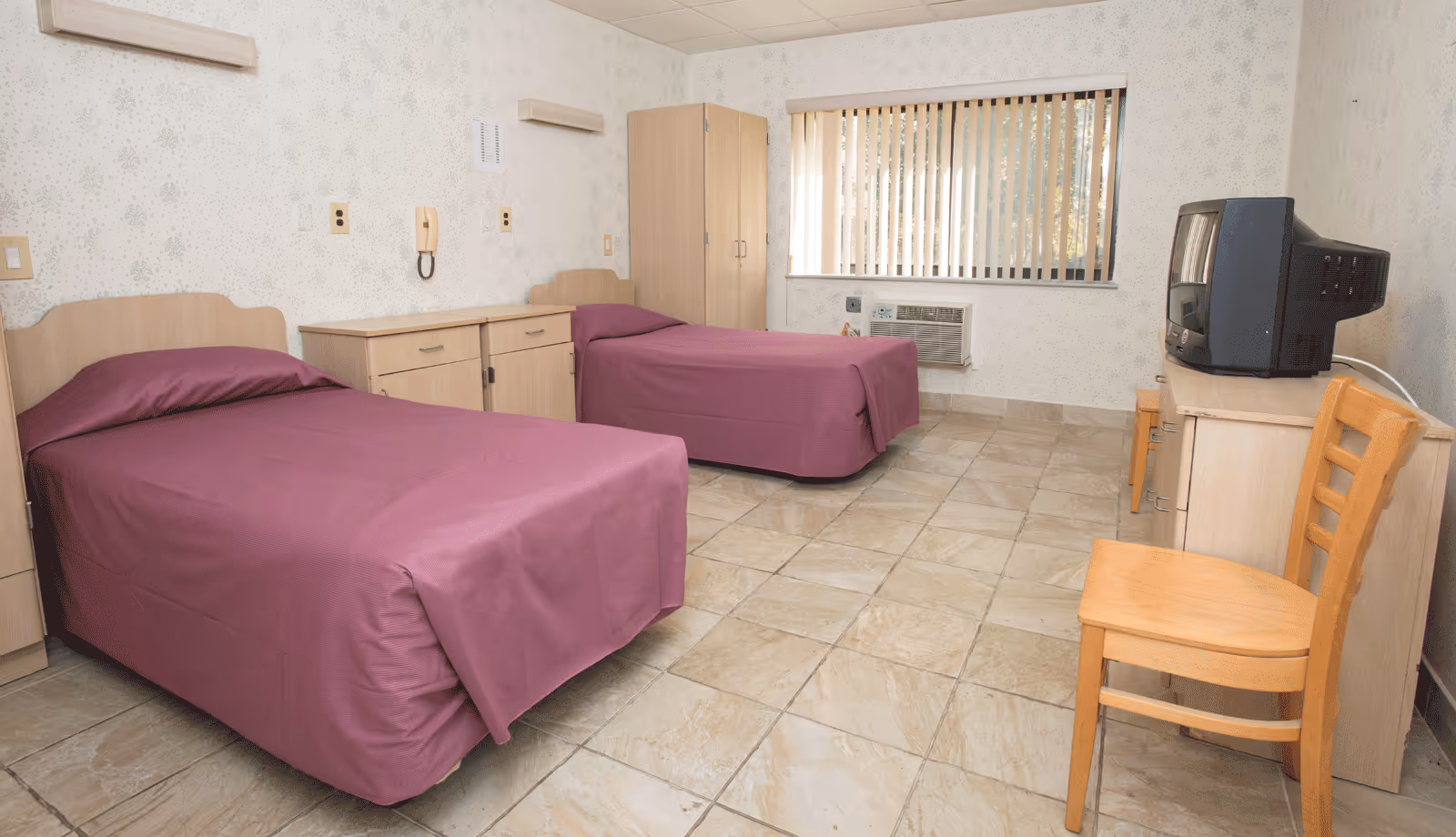 A simple bedroom with two single beds covered in purple bedspreads, a wooden chair, a small wooden dresser with an old-style CRT television on top, a wardrobe, and a window with vertical blinds. The floor is tiled and the walls have a light patterned wallpaper.