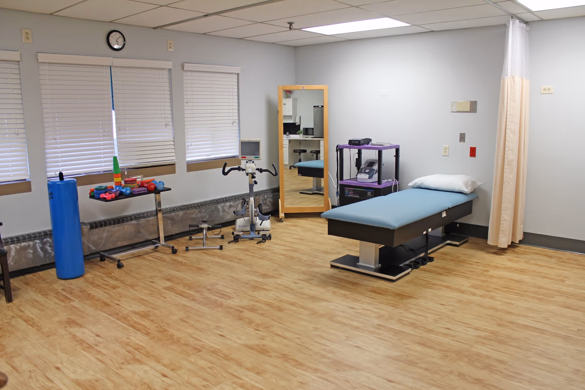 A physical therapy or rehabilitation room with a padded treatment table, exercise equipment including hand weights, a stationary bike, a large mirror, and a curtain divider. The room has wood-patterned flooring, white walls, and three windows with closed blinds.