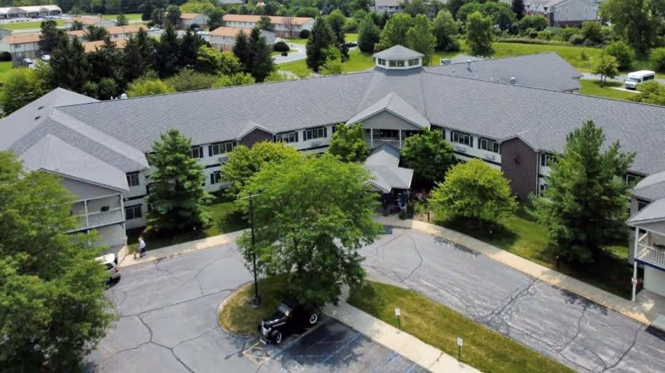 Aerial view of a large senior living community building with a gray roof surrounded by trees and greenery. There is a circular driveway with parked cars and a small grassy island with a tree in the center. Residential houses and more greenery are visible in the background.