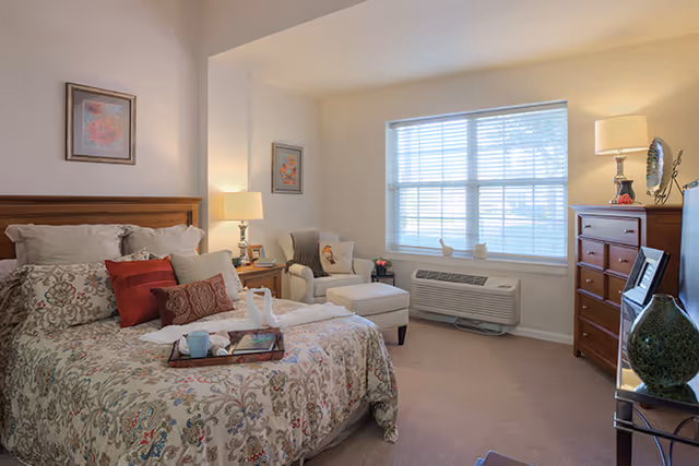 Bright, tastefully decorated bedroom with a patterned bed, an upholstered armchair and ottoman, wooden dresser, and a large window with blinds.
