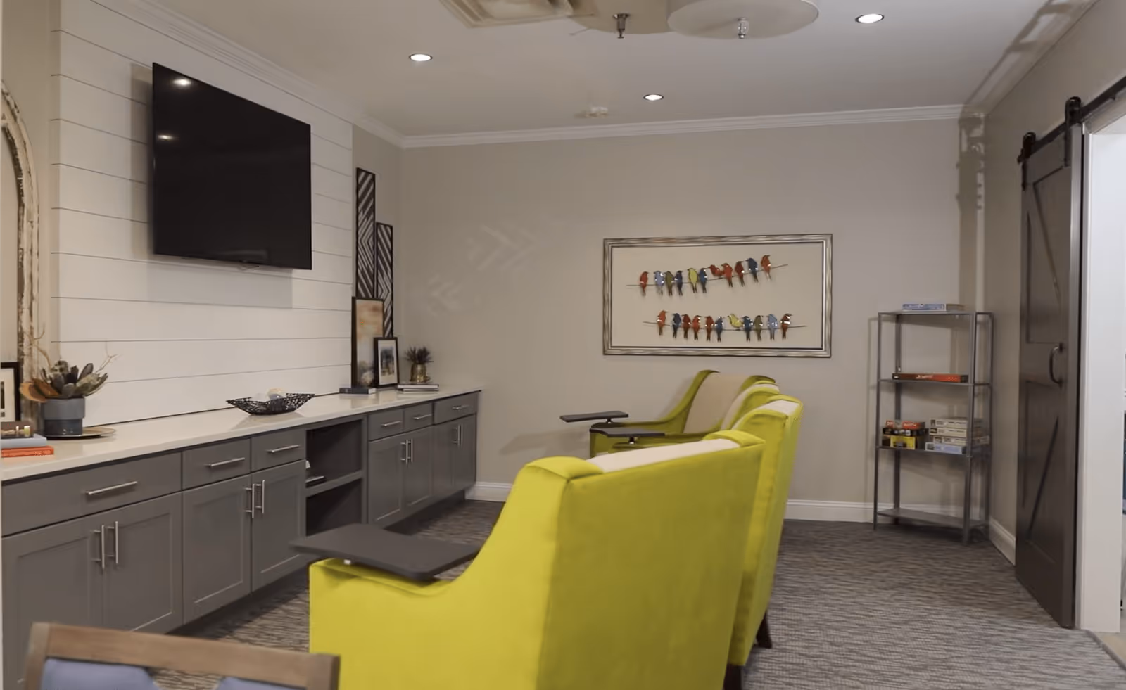 A cozy interior room with three bright green armchairs equipped with small attached tables facing a wall-mounted flat screen TV. The room features gray cabinetry with decorative items on top, a framed artwork of colorful birds on the wall, a metal shelving unit with board games, and a sliding barn door on the right side.