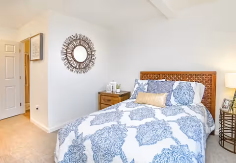 A bedroom with a bed featuring a wooden headboard and blue and white patterned bedding. There are several pillows on the bed, a wooden nightstand with decorative items, a round mirror with a sunburst frame on the wall, and a lamp on a small round table next to the bed. The room has light-colored walls and carpeted flooring.