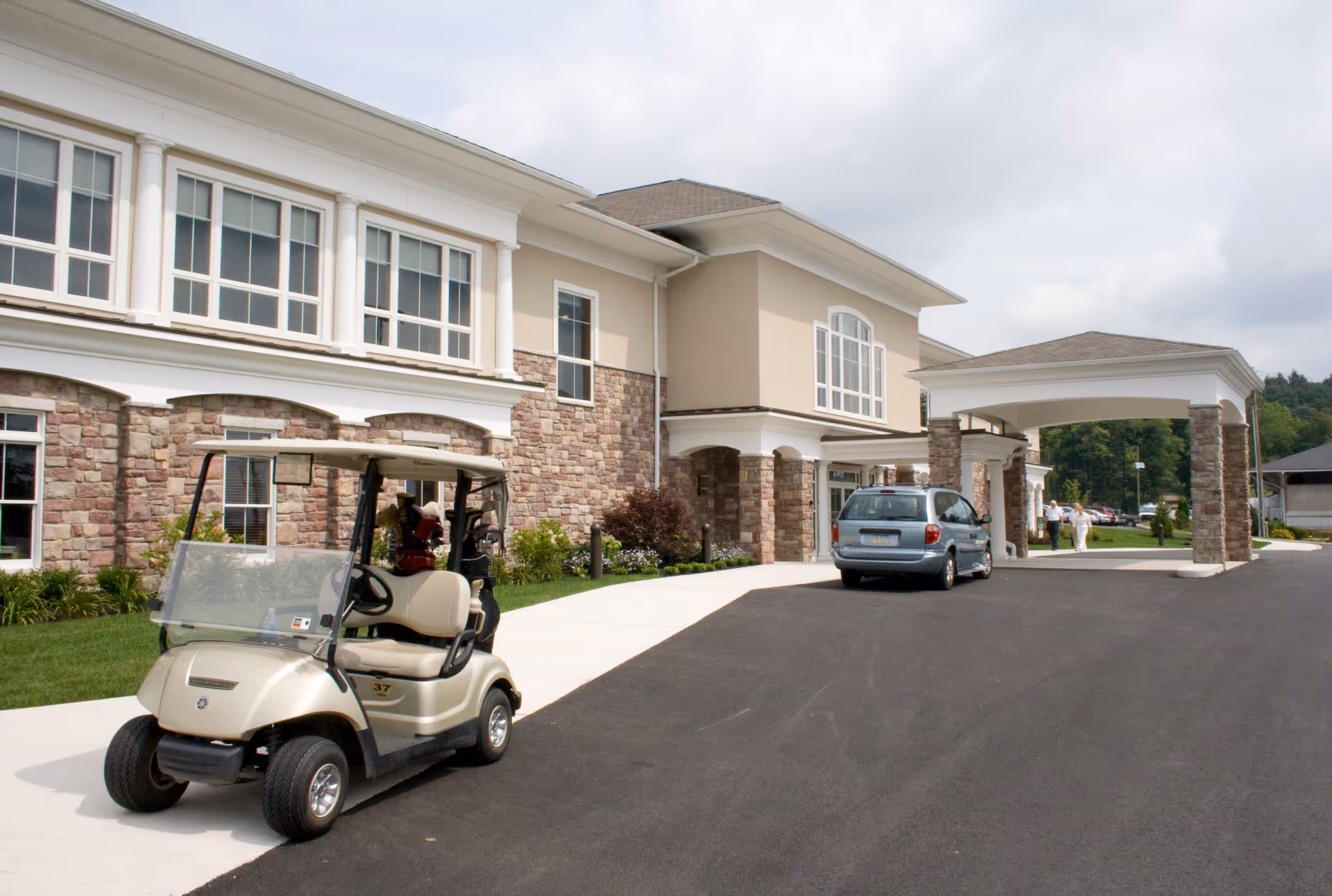 Front entrance of a senior living facility showing a porte-cochere with a parked golf cart and a car on the driveway.