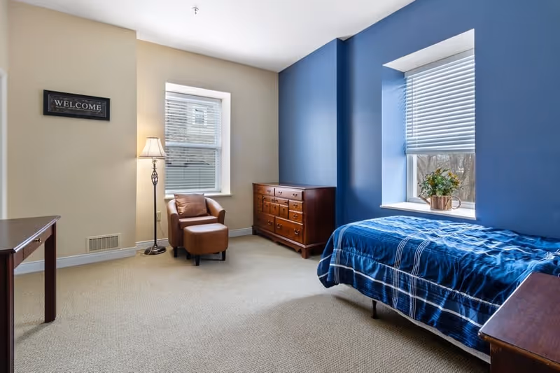 A bright and cozy bedroom with beige and blue walls, featuring a single bed with blue plaid bedding, a wooden dresser, a brown armchair with an ottoman, a floor lamp, and two windows with blinds. A small plant sits on the windowsill, and a 'WELCOME' sign is mounted on the beige wall.