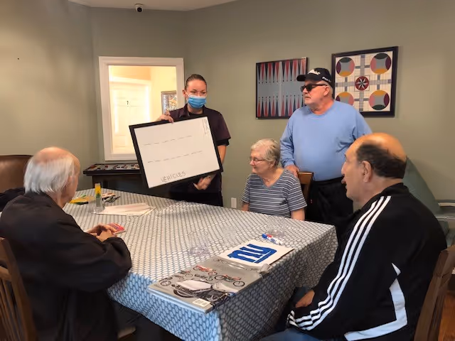 A group of four elderly people sitting around a table in a room, with a staff member wearing a mask standing and holding a whiteboard labeled 'VEHICLES'. The table is covered with a patterned tablecloth and some papers and markers are on it. The room has light-colored walls with two framed artworks hanging.