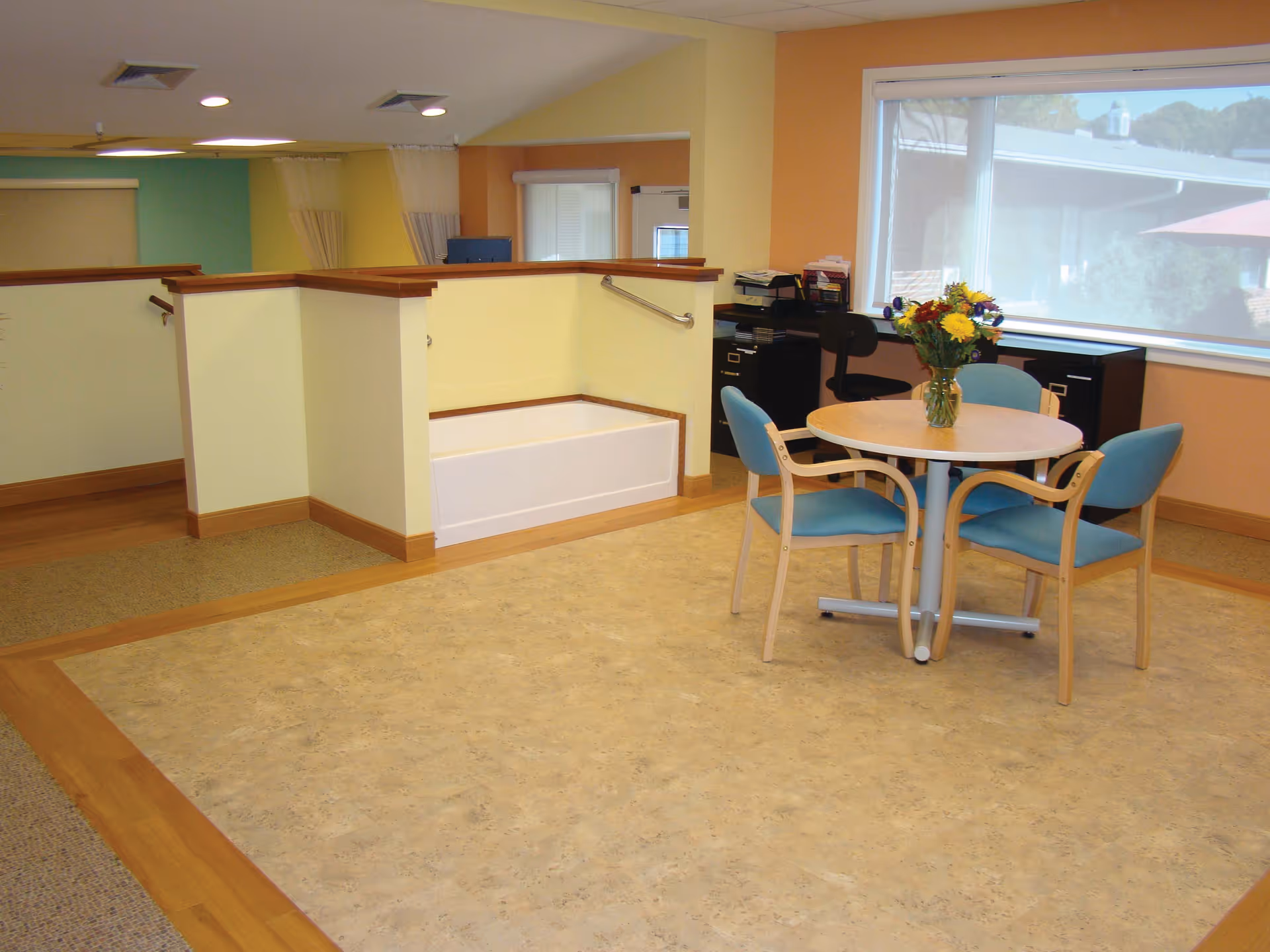 A bright interior room with a round table and four blue cushioned chairs. A vase with colorful flowers is placed on the table. The room has large windows with a view outside, light yellow walls, and a small built-in bench area with handrails. There are office desks and chairs near the window.