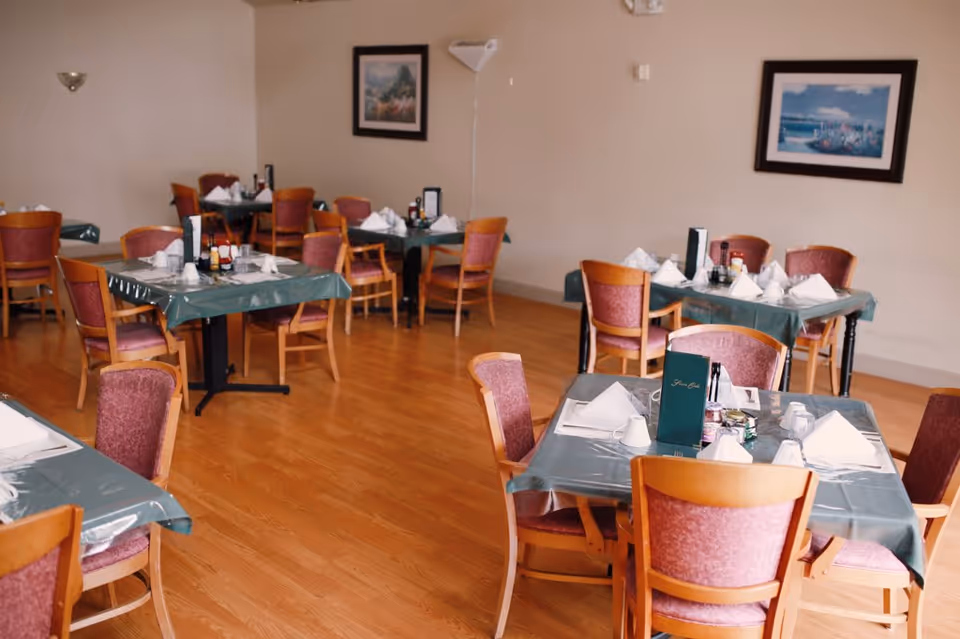 A dining room with multiple tables covered in green tablecloths, place settings, menus, and wooden chairs on a hardwood floor.
