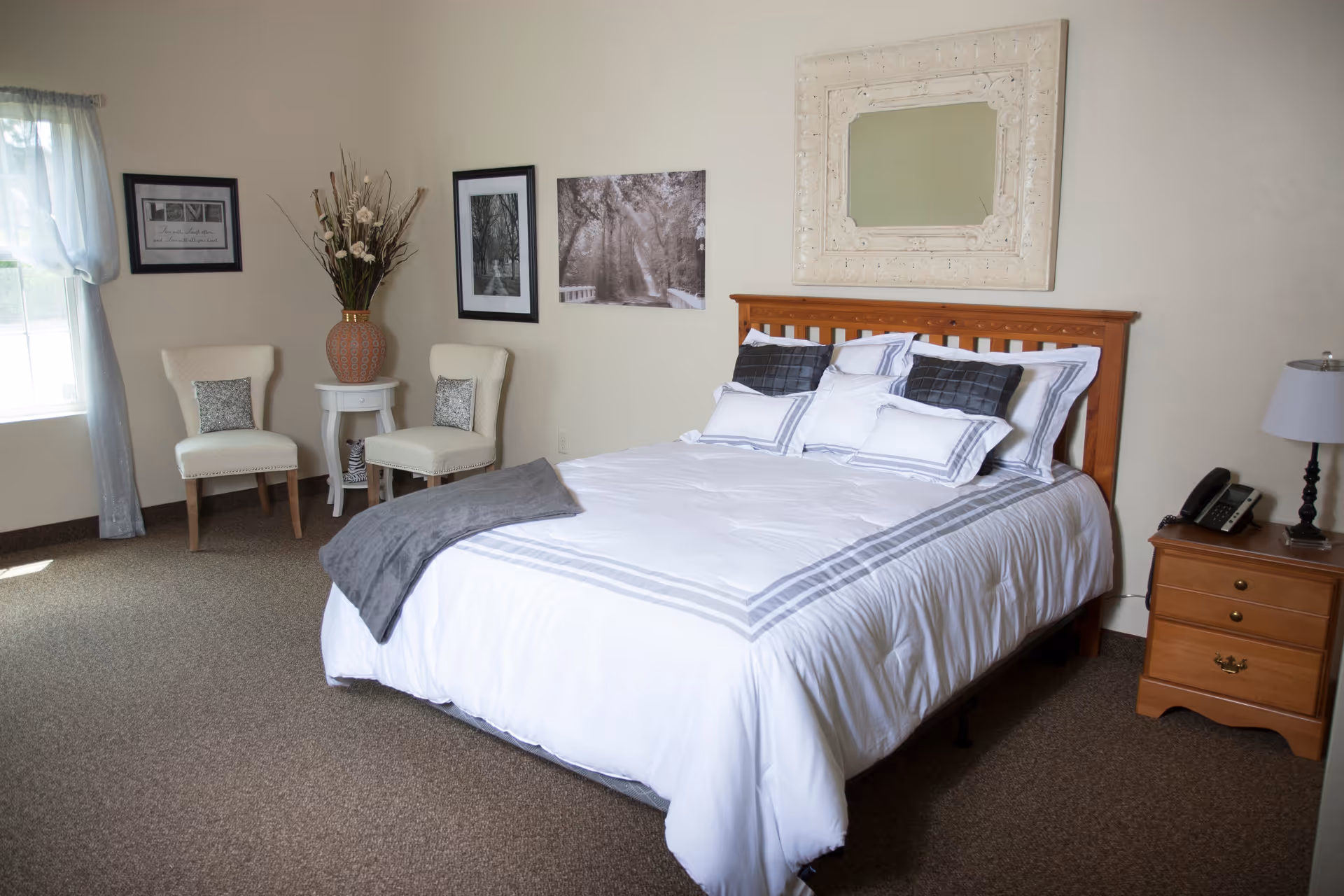 Bright, well-decorated bedroom with a made bed, nightstand, two chairs, and wall art.