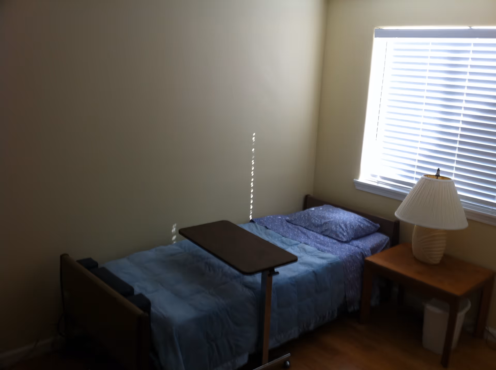 A small bedroom with a single bed covered in a blue blanket and a blue pillow. There is an overbed table positioned over the bed. Next to the bed is a wooden nightstand with a white lamp and a small trash bin underneath. A window with closed blinds is on the wall behind the nightstand.