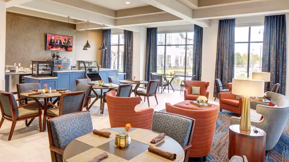 A bright and spacious dining and lounge area with multiple round tables and chairs arranged neatly. The room features large windows with blue curtains allowing natural light to fill the space. There are comfortable armchairs and ottomans in shades of orange and gray, a small counter with a coffee machine and display case, and a wall-mounted TV showing a talk show.