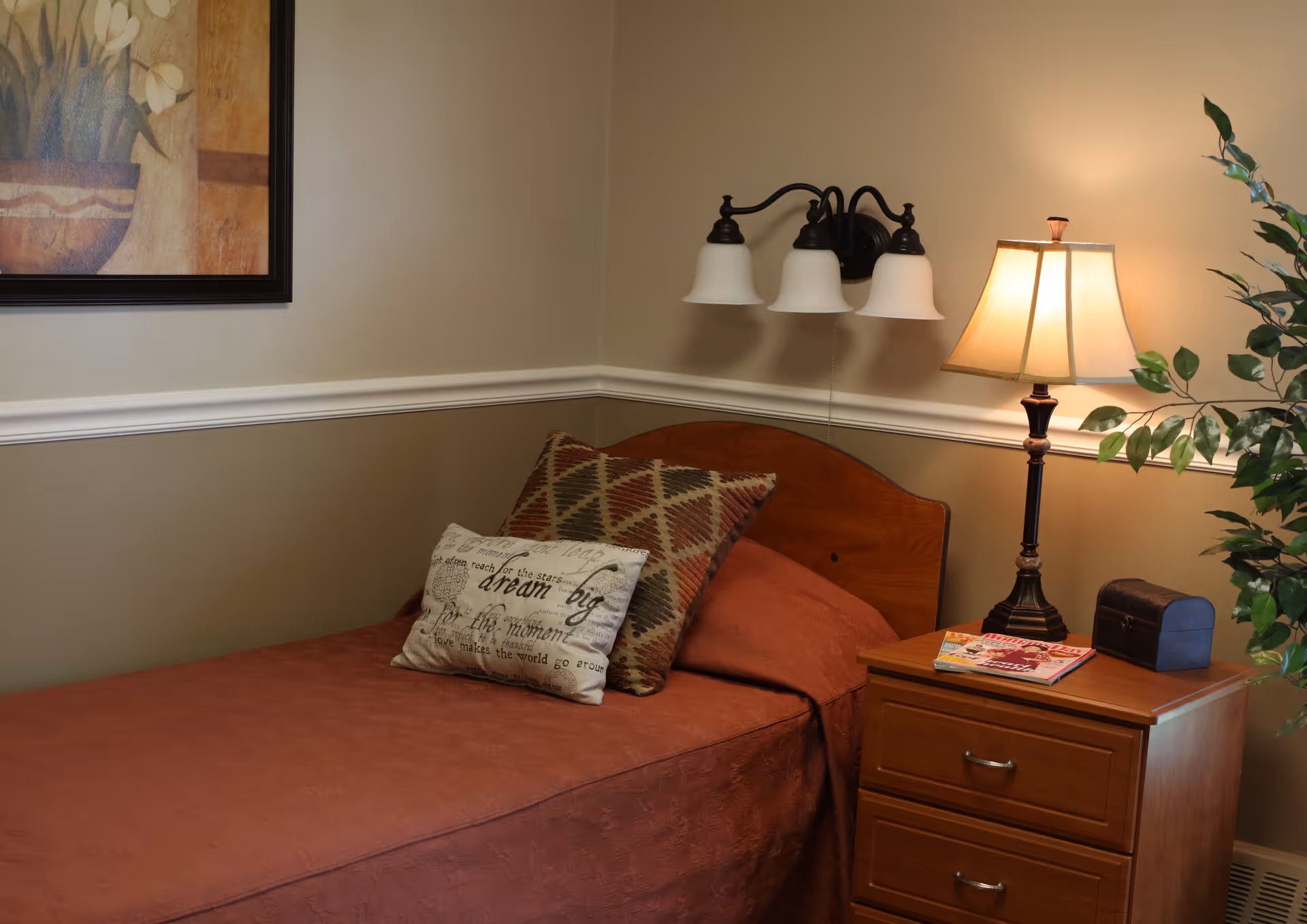A cozy bedroom with a single bed covered in a rust-colored bedspread, two decorative pillows, a wooden headboard, a nightstand with a lamp, a small chest, and magazines. A wall-mounted light fixture and a framed floral painting are visible on the beige walls, along with a green potted plant beside the nightstand.
