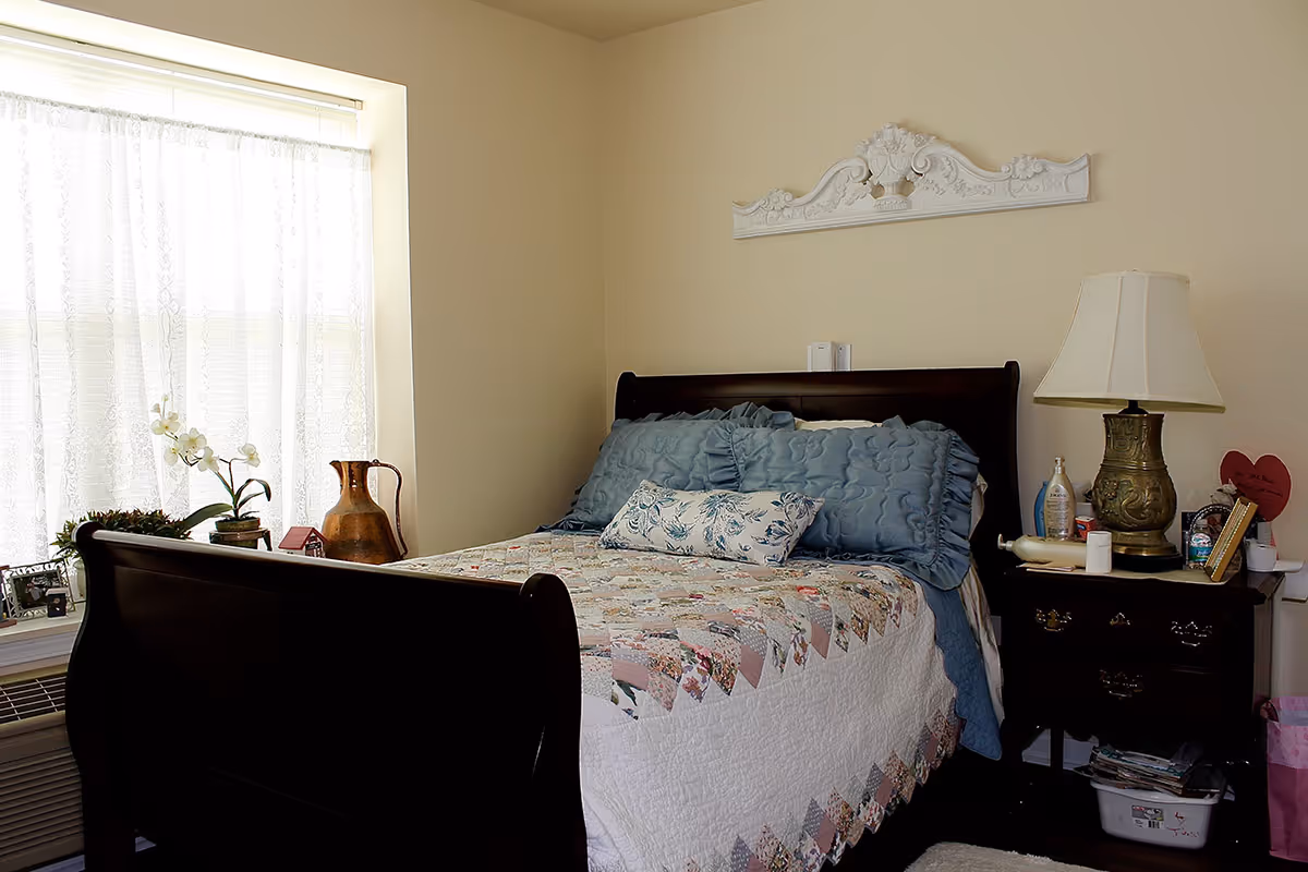 A sunlit bedroom featuring a dark wooden sleigh bed with a patchwork quilt and pillows, a nightstand with a lamp and decor, and a window with lace curtains.