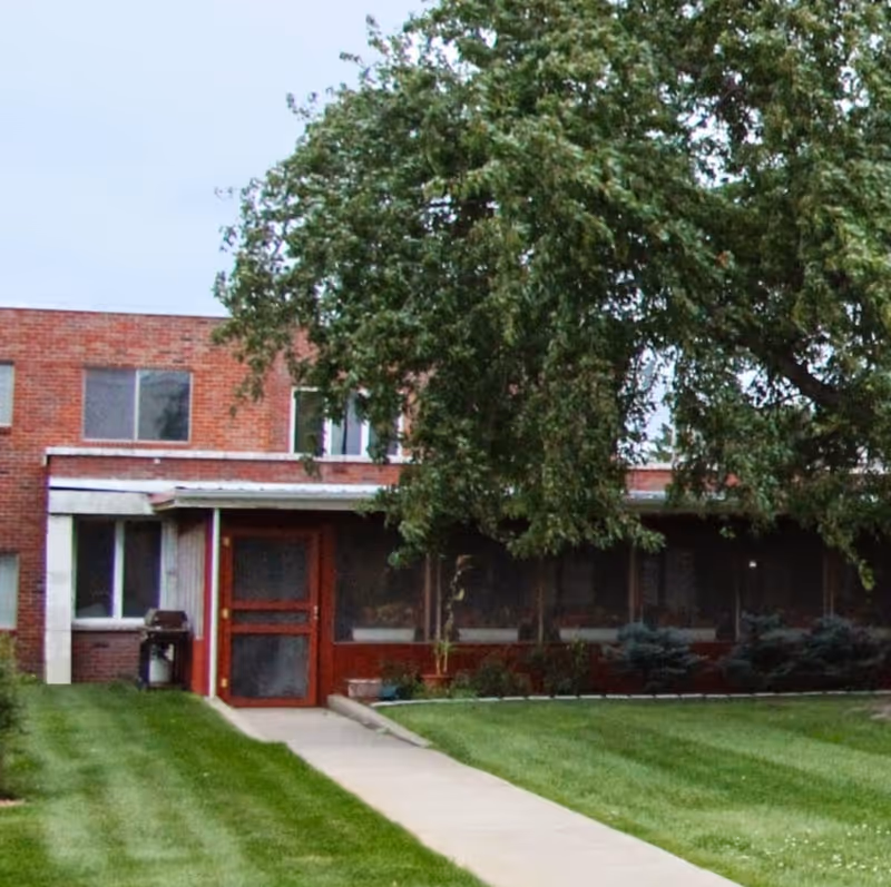 Brick building front with a walkway leading to a screened entrance, a large overhanging tree and a manicured lawn.