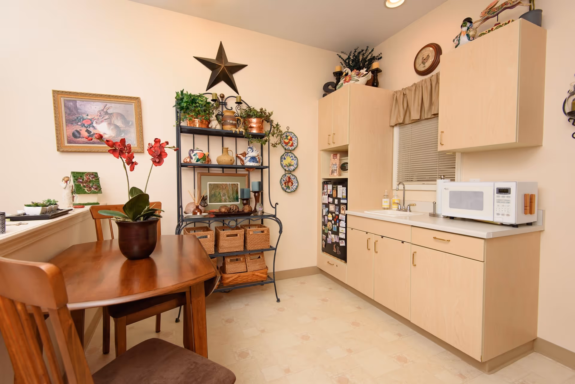 A cozy kitchen area with light wood cabinets, a white countertop, a microwave, and a small sink under a window with blinds and a beige valance. To the left, there is a wooden dining table with two chairs and a potted plant with red flowers on the table. A black metal shelving unit holds decorative items, plants, and baskets. The walls are light-colored and decorated with a framed picture of rabbits, a large star, and three decorative plates.