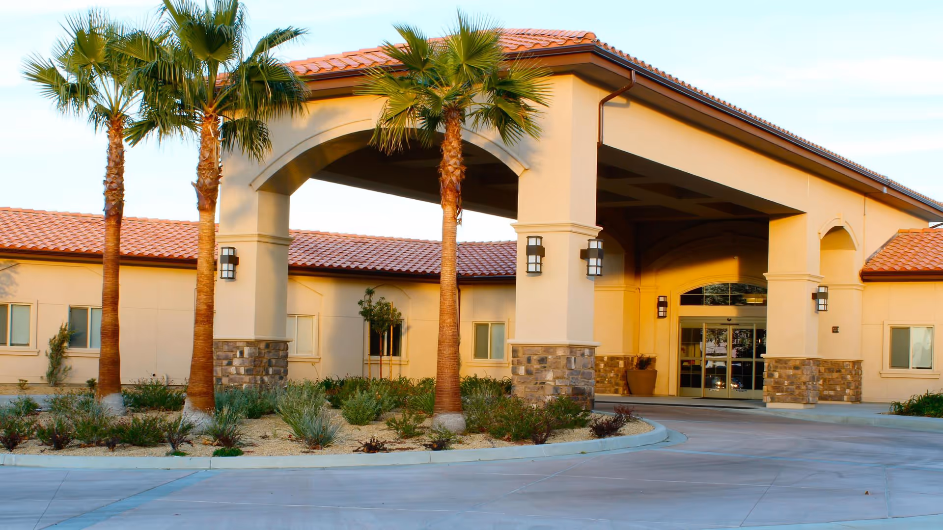 Exterior view of The Ellison John Transitional Care Center featuring a covered entrance with stone and stucco pillars, a tiled roof, palm trees, and landscaped plants in front.