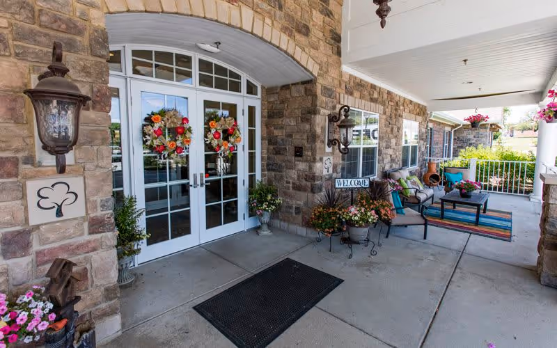 Entrance area of Bickford of Greenwood featuring double glass doors decorated with floral wreaths, stone walls, outdoor seating with chairs and a table on a colorful rug, potted plants, and a welcome sign under a covered porch.