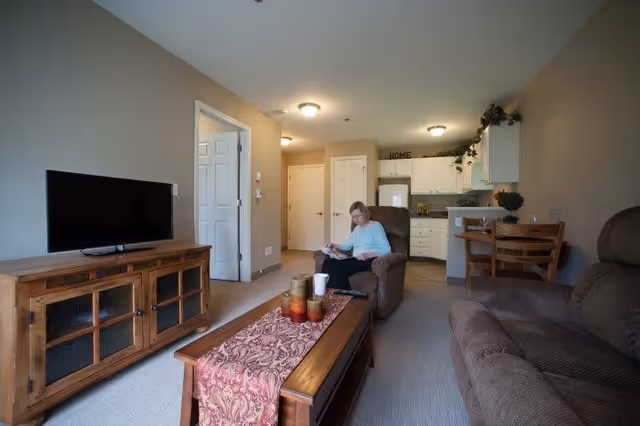 A cozy living room area in a senior living facility with a woman sitting in a recliner chair reading a book. The room features a wooden TV stand with a flat-screen TV, a wooden coffee table with decorative candles and a runner, a brown sofa, and a small dining table with chairs near a kitchen area with white cabinets and appliances. The walls are painted beige, and there are ceiling lights illuminating the space.