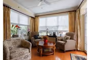 A cozy living room with large windows covered by white blinds and gold curtains. The room features two beige armchairs, a patterned armchair, a round wooden coffee table with a red flower centerpiece, and a ceiling fan above. There is a small plant on the windowsill and a view of the outdoors through the windows.