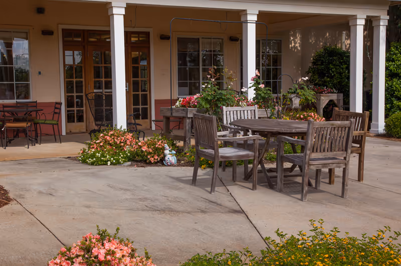 Outdoor patio area at The Brennity at Daphne Assisted Living & Memory Care featuring a round wooden table with four wooden chairs on a concrete surface, surrounded by flower beds with pink and yellow flowers. The patio is adjacent to a building with large windows and a covered porch supported by white columns.