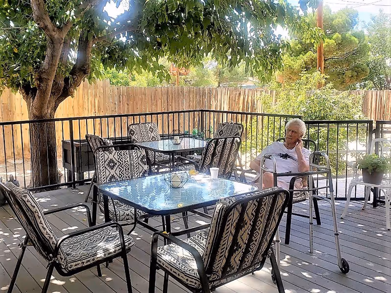 An elderly person sitting on a chair with a walker next to them on a shaded outdoor patio. The patio has several glass-top tables and cushioned chairs with patterned fabric. There is a wooden fence and trees in the background providing shade.