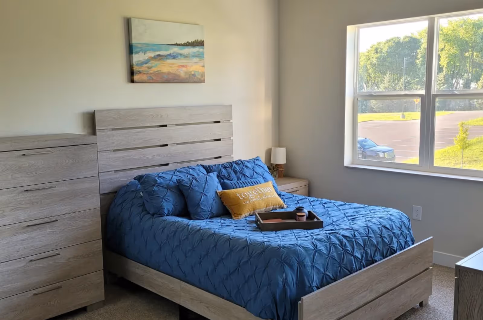 A bedroom with a wooden bed frame and matching dresser. The bed is made with a blue comforter and several blue pillows, along with a yellow decorative pillow that says 'together is our favorite place to be.' There is a wooden tray with items on the bed. A window on the right side lets in natural light, showing a view of trees and a parked car outside. A painting of a beach scene hangs on the wall above the bed.