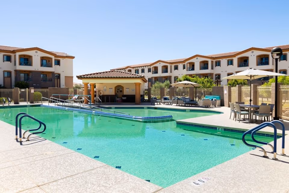 Outdoor swimming pool area at Fellowship Square Surprise with clear blue water, poolside tables with umbrellas, lounge chairs, and a small shaded pavilion. The background shows a multi-story residential building under a clear blue sky.