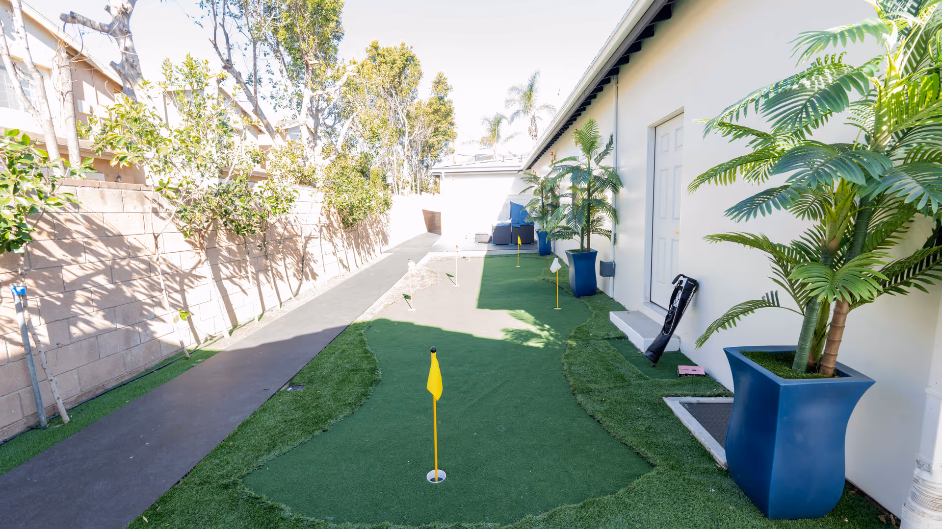 Outdoor putting green area with several small golf holes marked by yellow flags, adjacent to a white building with potted plants in blue planters and a paved walkway alongside a brick wall with trees.