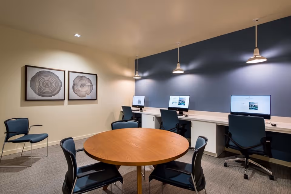 A modern computer room with a round wooden table surrounded by four chairs in the foreground. Against the back wall, there are three computer workstations with monitors and office chairs, illuminated by three pendant lights. The walls are painted in light beige and blue, with two framed artworks depicting tree rings on the left wall.