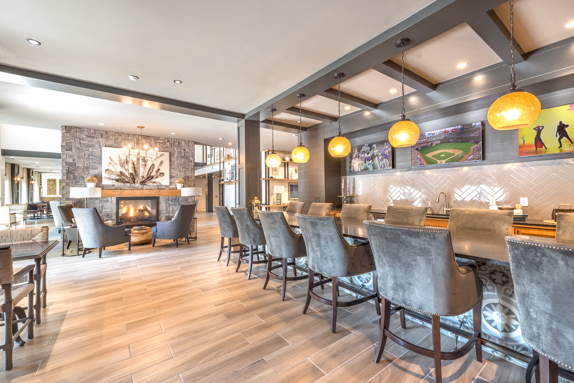 A spacious and modern common area in a senior living facility featuring a long bar counter with high-backed chairs, hanging pendant lights, and three wall-mounted televisions displaying sports and dance images. To the left, there is a cozy seating area with gray armchairs arranged around a stone fireplace with a feather artwork above it. The room has light wood flooring and a bright, open layout.
