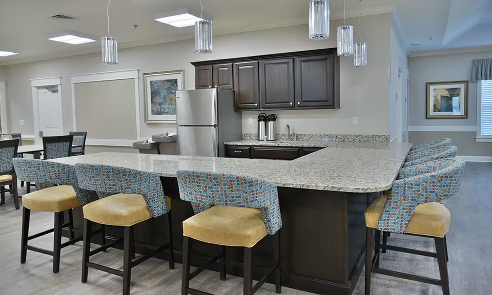 Communal kitchen with a large granite island surrounded by patterned barstools, dark cabinets, and a stainless steel refrigerator.