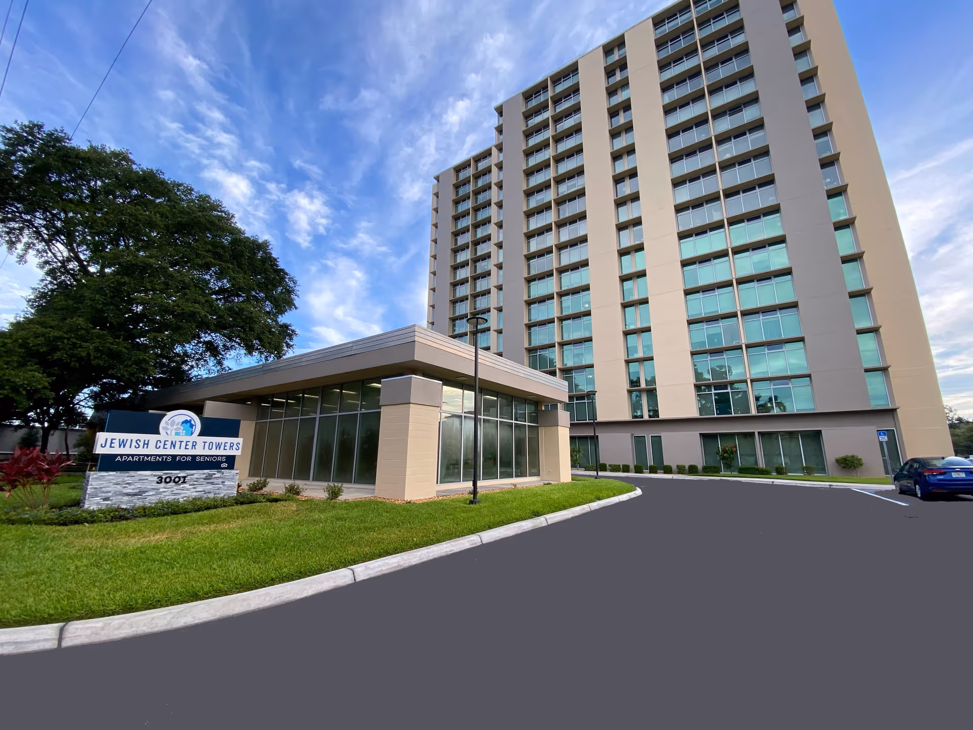 Exterior view of a tall senior living apartment building named Jewish Center Towers with a sign in front that reads 'Jewish Center Towers Apartments for Seniors'. The building is surrounded by a driveway, green grass, and trees under a partly cloudy blue sky.