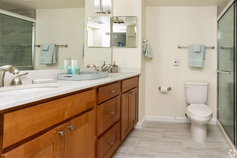 A clean and modern bathroom with a wooden vanity featuring two sinks and silver faucets. Above the vanity are two square mirrors and a wall-mounted light fixture. To the right, there is a white toilet with a towel rack above it holding a folded blue towel. Next to the toilet is a glass-enclosed shower with tiled walls. The floor is covered with light-colored tiles.
