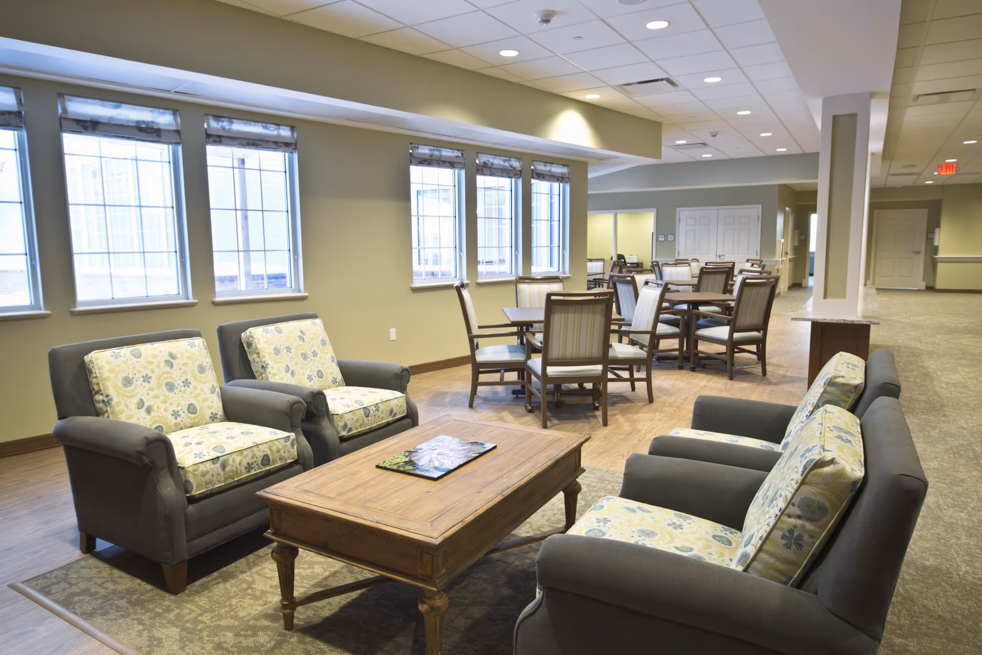 A spacious and well-lit common area with multiple armchairs featuring floral cushions arranged around a wooden coffee table. In the background, there are several dining tables with chairs. Large windows line one wall, allowing natural light to fill the room. The flooring is wood with a carpet under the seating area, and the walls are painted a soft green.
