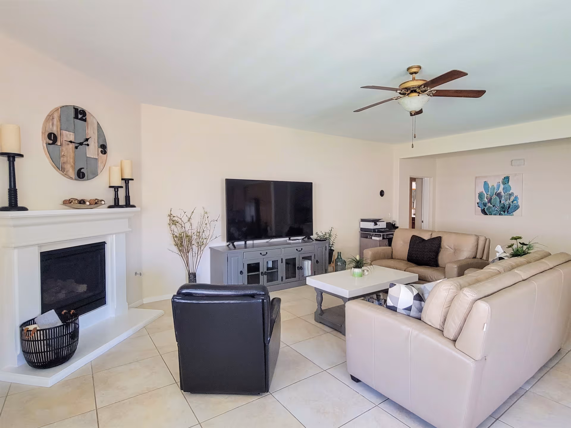 A bright living room with beige leather sofas, a black armchair, a white coffee table, a large flat-screen TV on a gray TV stand, a white fireplace with candles and a decorative clock above it, a ceiling fan with wooden blades, and a painting of a cactus on the wall.