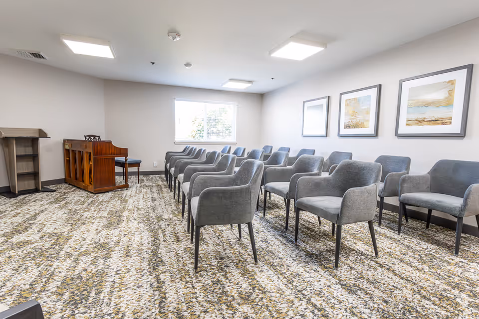 A bright room with rows of gray upholstered chairs arranged facing a wooden piano and a lectern. The room has a patterned carpet, white walls with three framed abstract paintings, and a window letting in natural light.