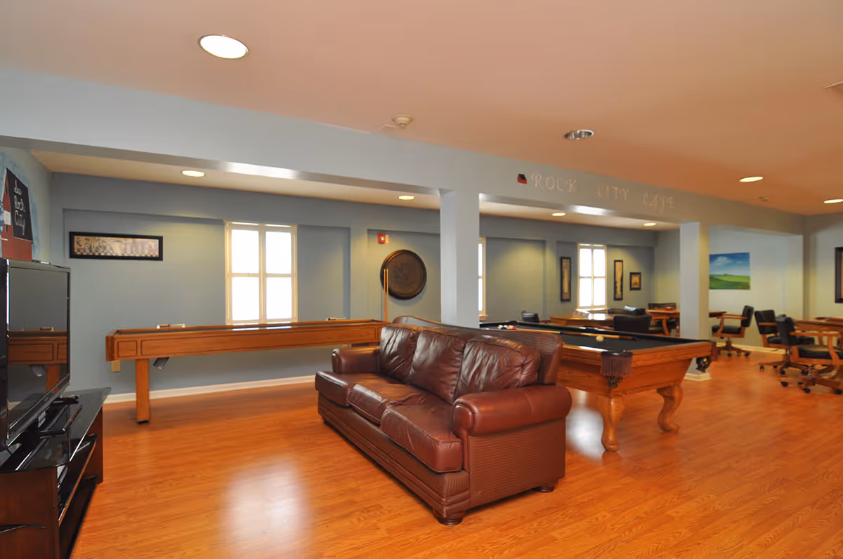 A spacious recreational room with wooden flooring featuring a brown leather sofa, a pool table, a shuffleboard table, and several tables with chairs in the background. The walls are painted light blue with windows letting in natural light. The ceiling has recessed lighting and a sign that reads 'Rock City Cafe'.