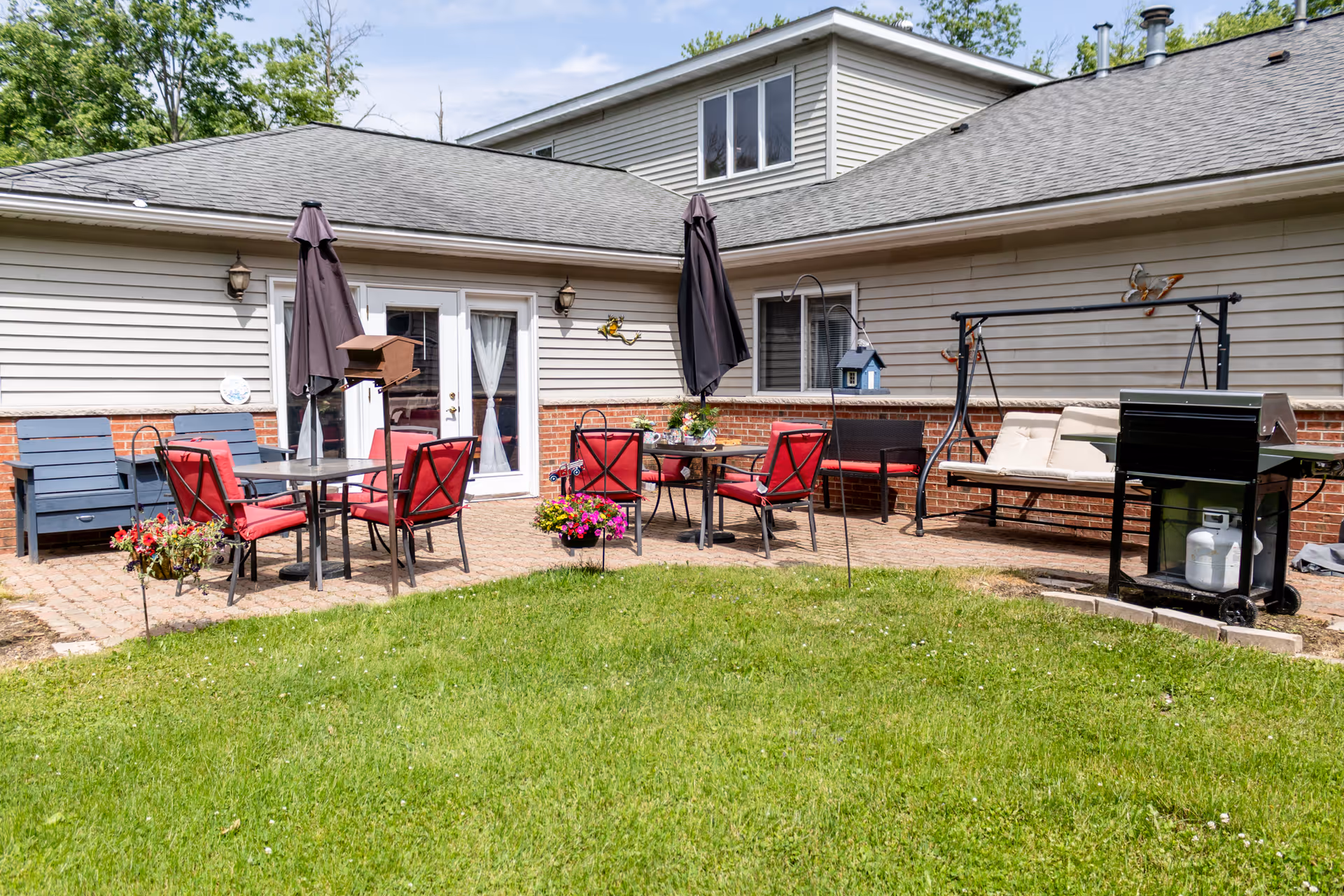Outdoor patio area with red cushioned chairs around tables, two closed black umbrellas, a swing bench with beige cushions, a black grill, and flower pots on a green lawn in front of a beige and brick building.